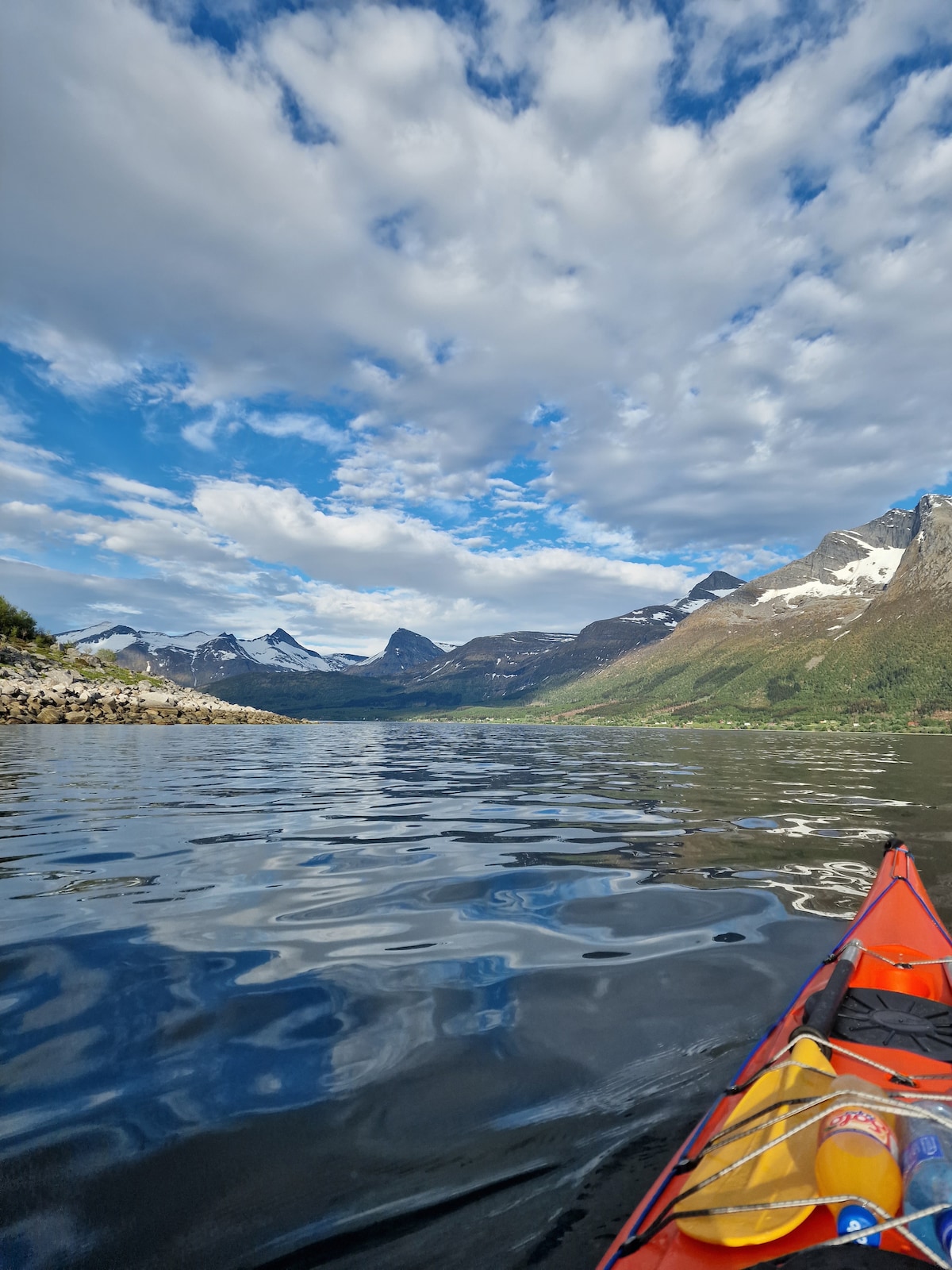 The view is captured from a kayak on calm waters, with ripples reflecting the sky above. Majestic mountains, partially adorned with snow, frame the landscape. A mix of green hills and rocky shoreline can be seen along the water's edge, under a backdrop of scattered clouds.