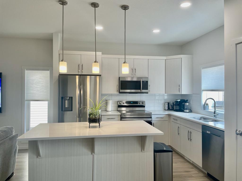 A modern kitchen is shown, featuring sleek white cabinetry and stainless-steel appliances. Three pendant lights hang over a central island, which offers additional countertop space. A sink is located near a window, allowing natural light to fill the area.