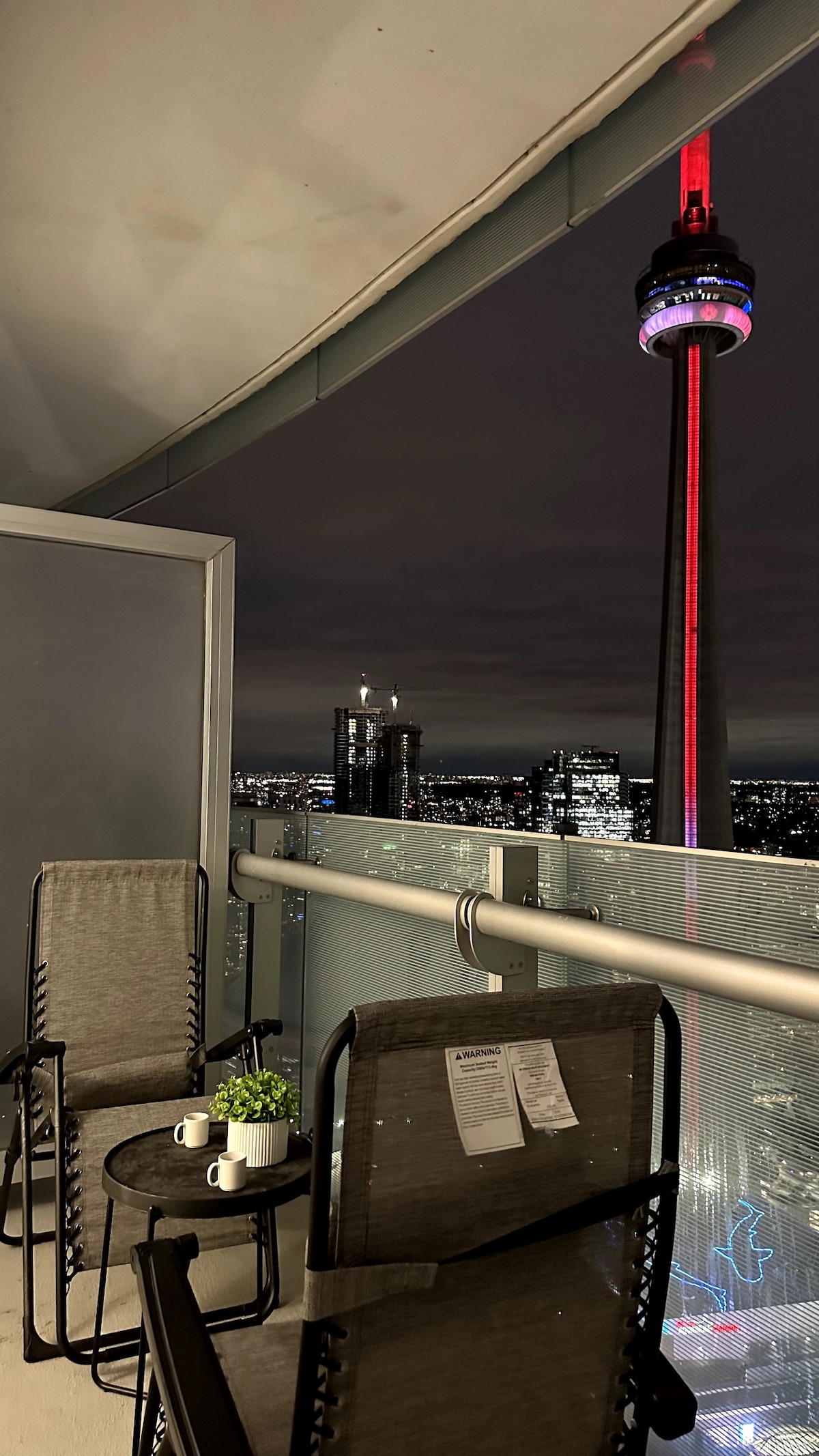 A private balcony offers two seated chairs beside a small table, presenting a direct view of the CN Tower illuminated in red. The city skyline is visible in the background, creating an open space ideal for relaxation.