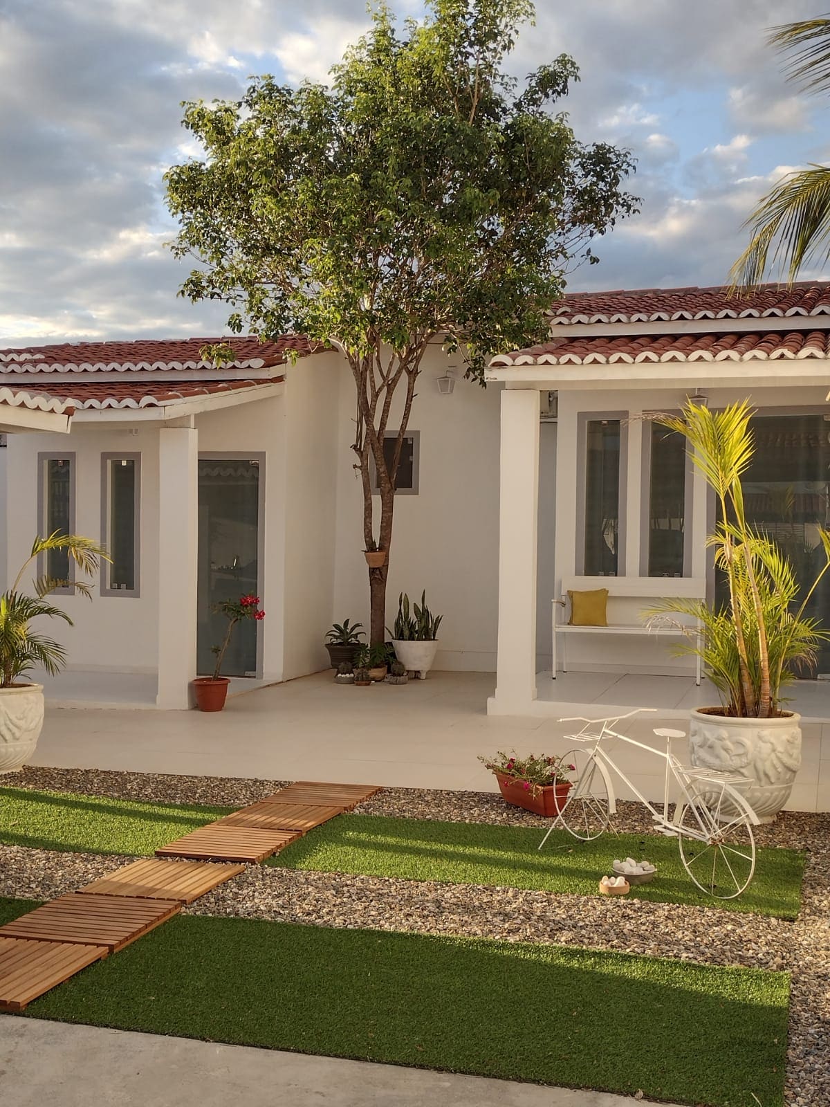 The exterior features a pathway made of wooden planks and gravel, surrounded by lush green grass. Potted plants and a small tree enhance the area. The house has white walls, large windows, and a red tiled roof, creating a clean and welcoming façade.