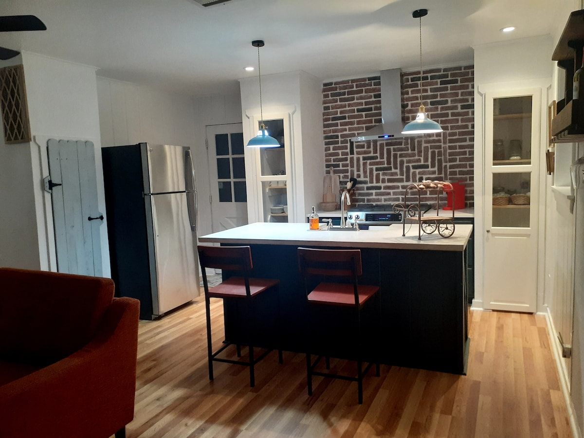 The kitchen features a spacious island with two bar stools, set against a striking brick backsplash. Stainless steel appliances include a refrigerator, complementing the modern design. Warm wood flooring contributes to a welcoming ambiance, and natural light filters in through the nearby windows.