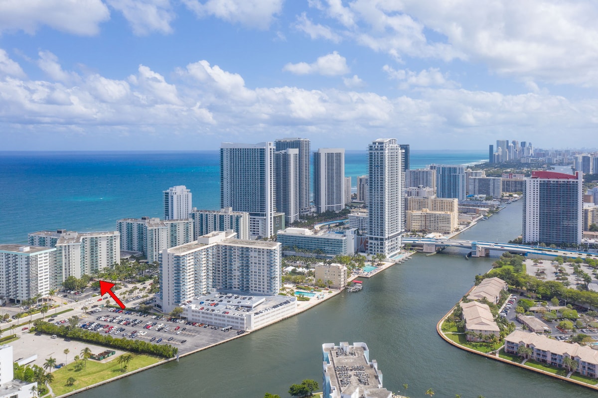 An aerial view captures a vibrant beachfront area, highlighting high-rise buildings along the coastline. The blue waters of the ocean contrast with the urban landscape, while the surrounding greenery and the city skyline form a picturesque backdrop.