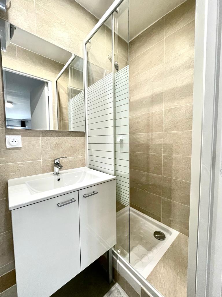 A modern bathroom features a sleek vanity with storage and a faucet. A glass shower enclosure is positioned beside the sink, showcasing neutral tiled walls. A large mirror enhances the bright space, reflecting natural light and creating an open feel.