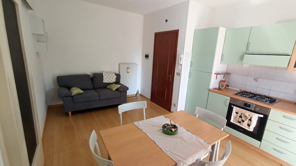 An open living space features a comfortable grey sofa adorned with green cushions. A wooden dining table is set with a centerpiece, while a light green kitchen with a stove is visible in the background. Natural light brightens the area, enhancing the inviting atmosphere.