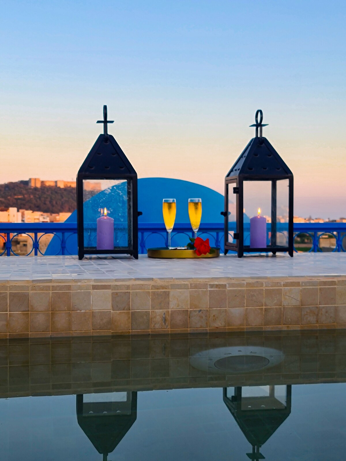 Two lanterns flank a tray with two flutes filled with a sparkling beverage, placed beside a serene water surface. A soft purple candle adds warmth, and a backdrop of a calm pastel sky is visible, creating a tranquil ambiance.