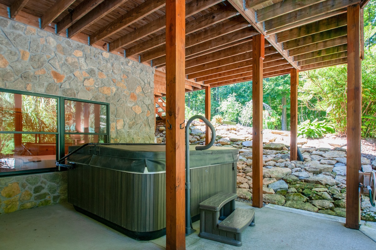A spacious patio area features a hot tub surrounded by natural stone walls and lush greenery. The wooden beams overhead provide structure, and large windows allow for glimpses into the inviting outdoor space. Steps lead up to the hot tub for easy access.