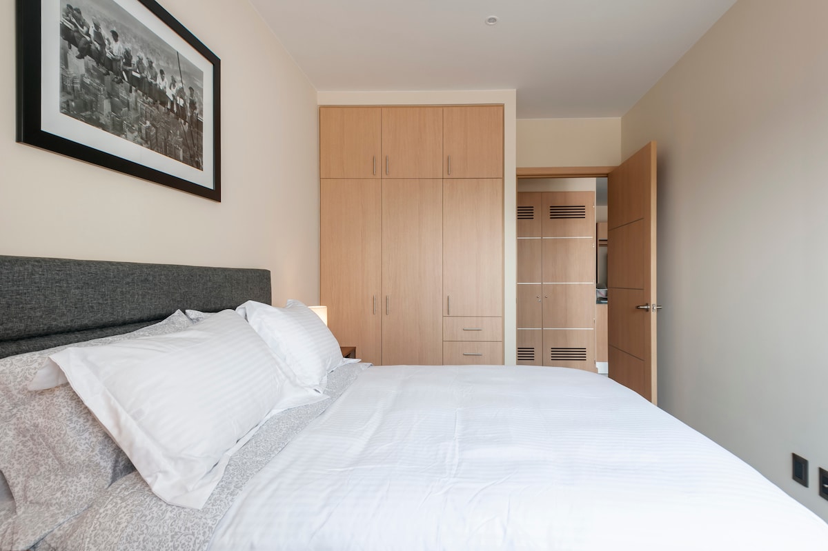A well-appointed bedroom features a spacious bed with white linens and a gray upholstered headboard. Natural light enters through a nearby window, illuminating the room. Light wood cabinetry lines the back wall, providing ample storage, while a framed black-and-white photo adds a decorative touch.