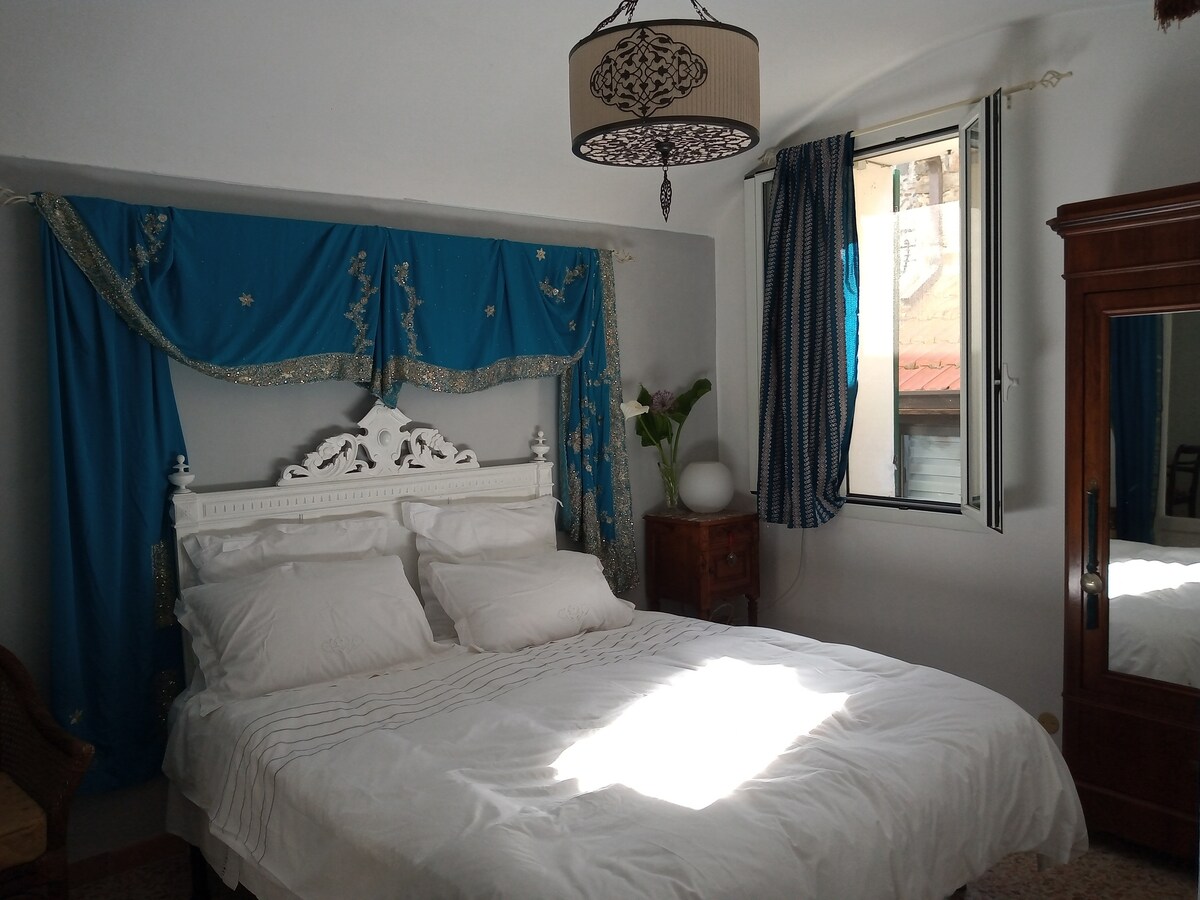 A spacious bedroom is illuminated by natural light from a window, with decorative blue draping and a stylish lamp overhead. The arrangement includes a large bed with plush white linens and a wooden wardrobe, enhancing the room's functional and inviting layout.