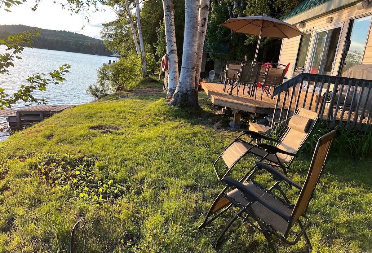 Lawn chairs are positioned on a grassy area near the water's edge, facing the lake. A wooden deck is visible nearby, furnished with a dining table and umbrella. The landscape includes birch trees and a view of the calm water, suggesting a serene outdoor setting.
