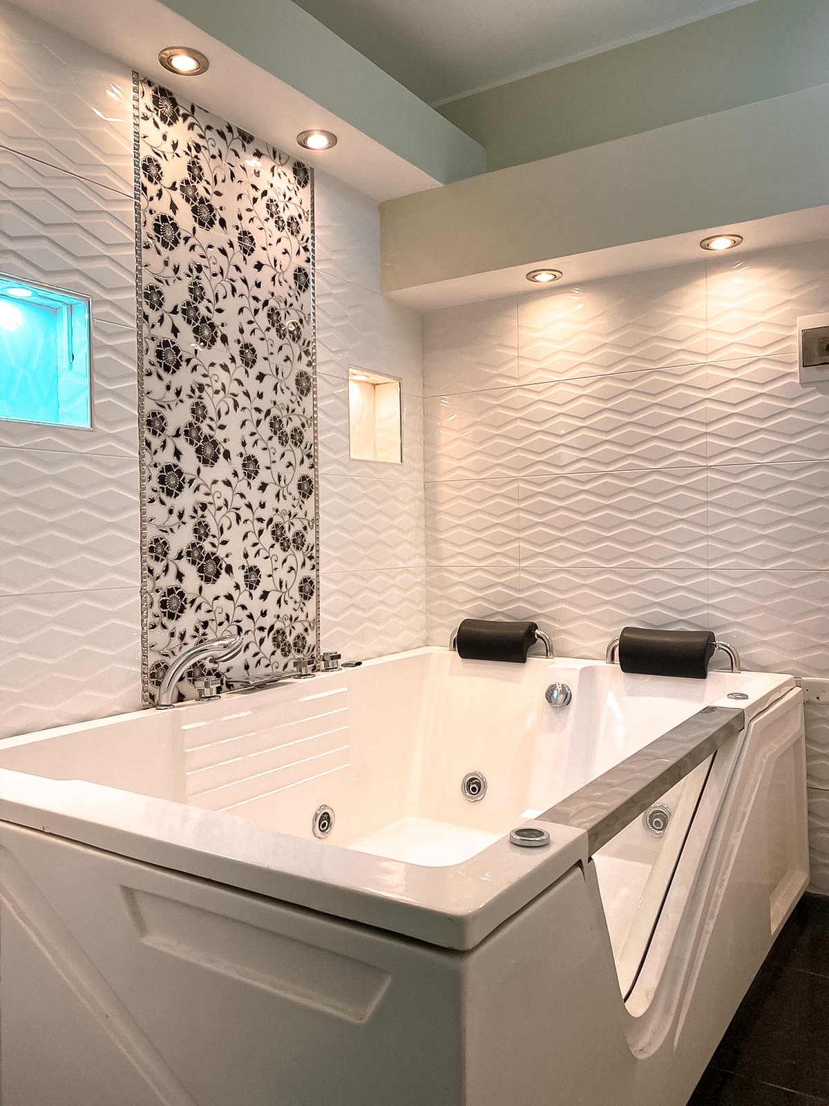 A modern jacuzzi is featured in a stylish bathroom, complemented by elegantly patterned wall tiles. Soft lighting highlights the sleek design, while two rolled towels are placed at either end of the jacuzzi for added comfort.