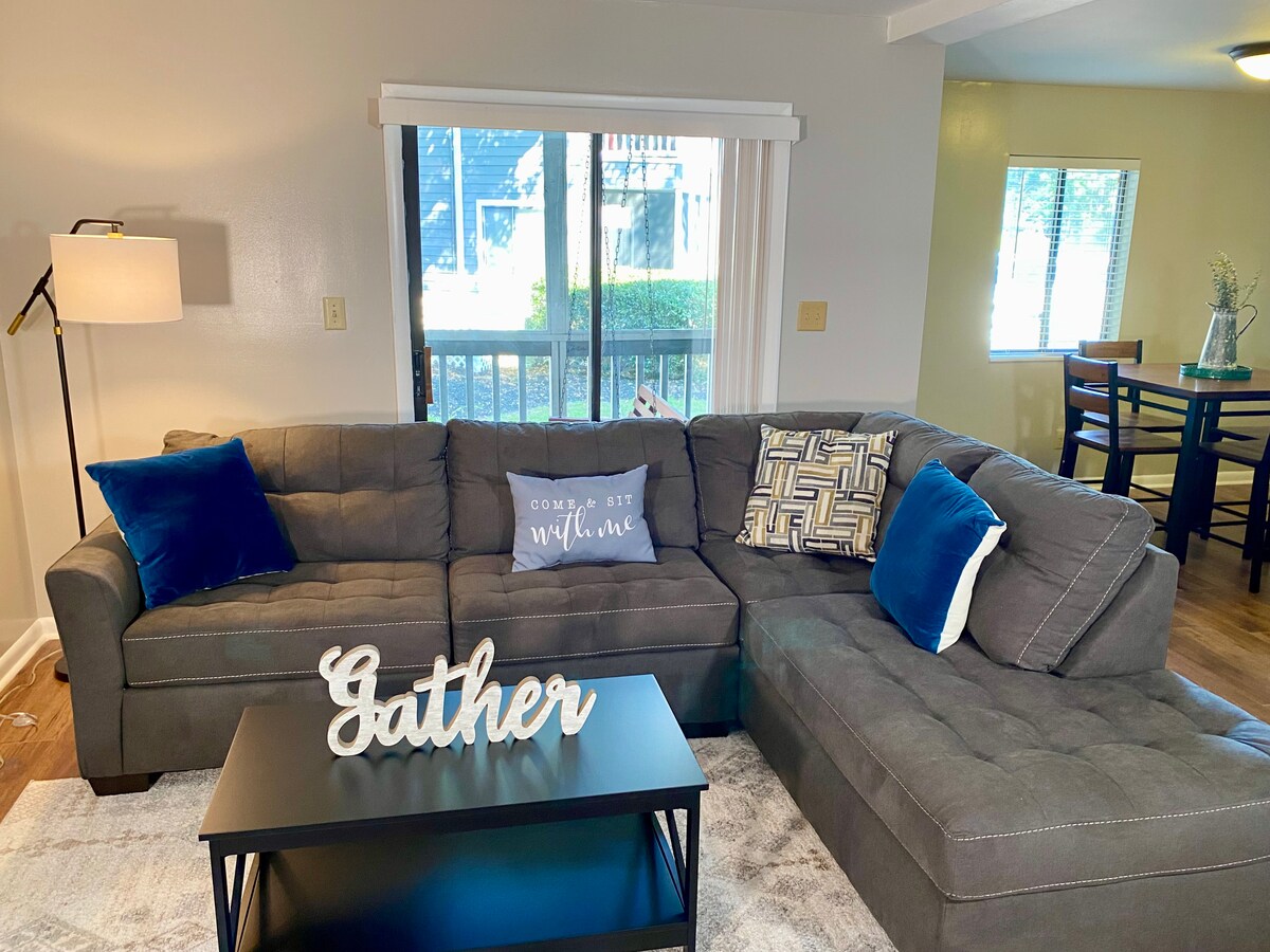A spacious living area features a gray sectional sofa adorned with decorative pillows in blue and gold tones. A black coffee table displays a wooden sign reading 'Gather.' Natural light floods in through large windows, illuminating the wood flooring and creating a welcoming atmosphere.