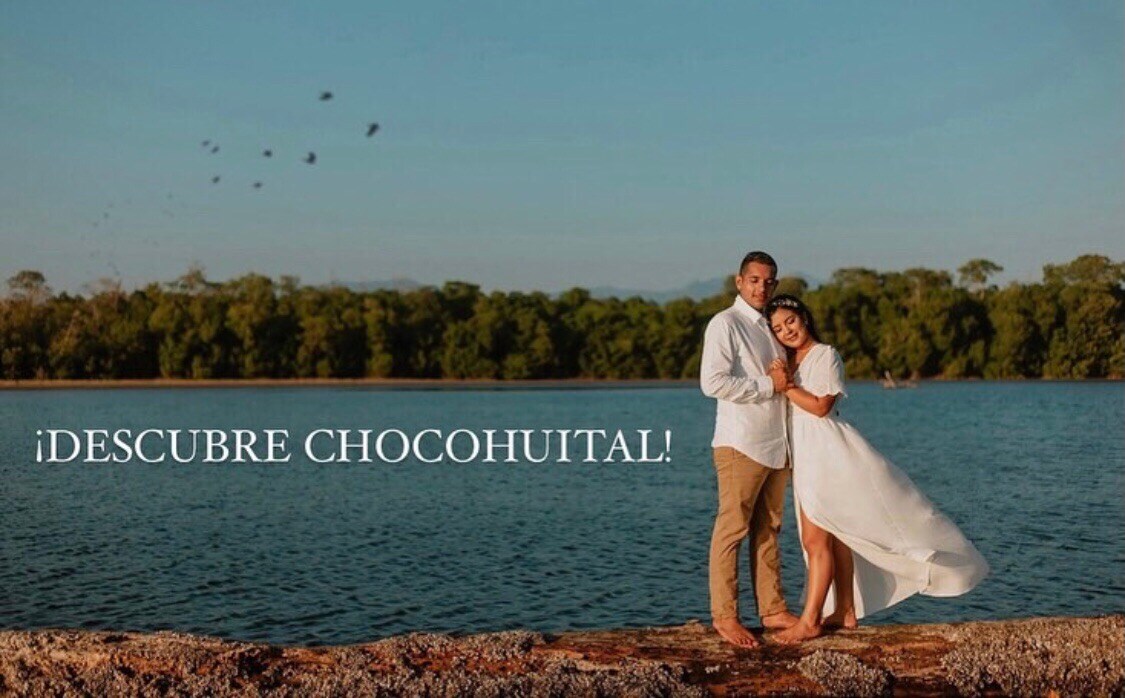 A couple is seen standing by a calm body of water, surrounded by lush greenery. The serene landscape features a sandy shoreline and a clear blue sky, with gentle waves lapping at the edge. Light clothing gently billows in the breeze, enhancing the tranquil atmosphere.