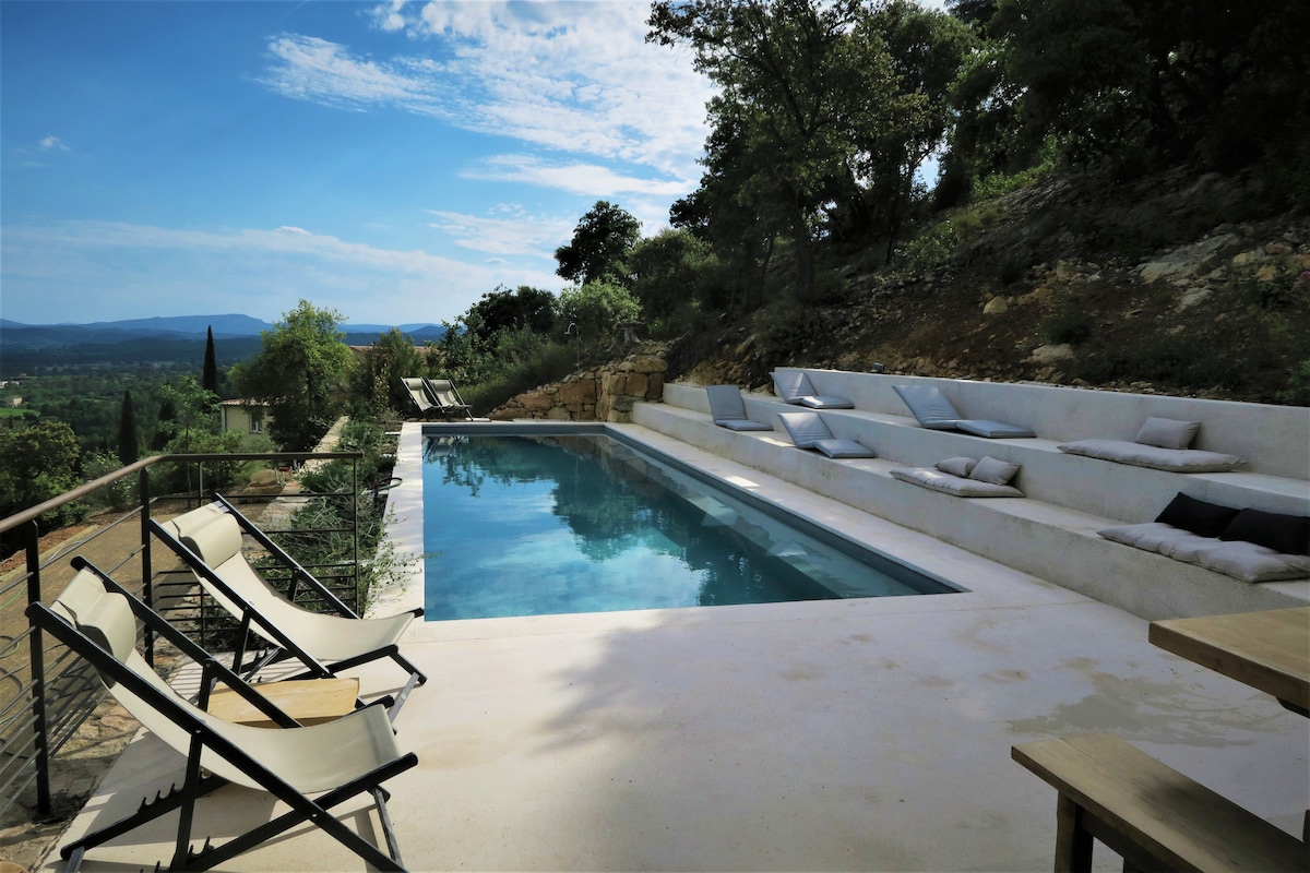 An outdoor pool area offers a serene view of the landscape, with a spacious swimming pool reflecting the sky. Comfortable lounge chairs are placed nearby. A natural stone wall and a variety of greenery surround the space, enhancing the tranquil setting.