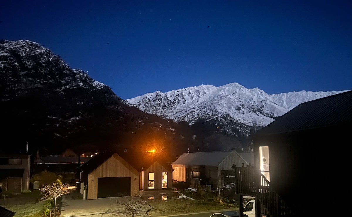 A serene evening view showcases snow-capped mountains under a night sky. Soft lights illuminate the nearby buildings, creating a welcoming ambiance. The landscape transitions from natural beauty to quiet residential structures, inviting tranquility and a sense of connection with the surroundings.