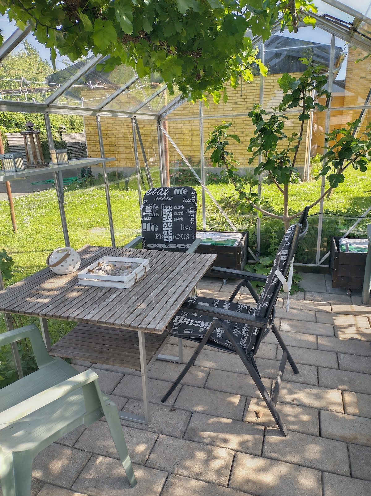 A greenhouse area is featured with a wooden table surrounded by black chairs. Potted plants are visible, adding greenery to the space. Outside, a well-maintained yard with grass can be seen, creating a tranquil setting for relaxation.