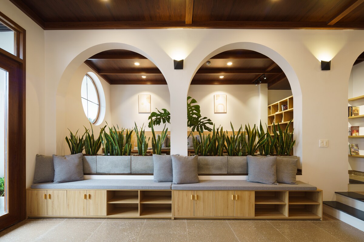 A spacious seating area features a long, built-in bench cushioned with soft gray pillows. Decorative green plants are arranged in planters, enhancing the serene environment. Arched windows provide natural light, while wooden accents in the ceiling create a warm backdrop.