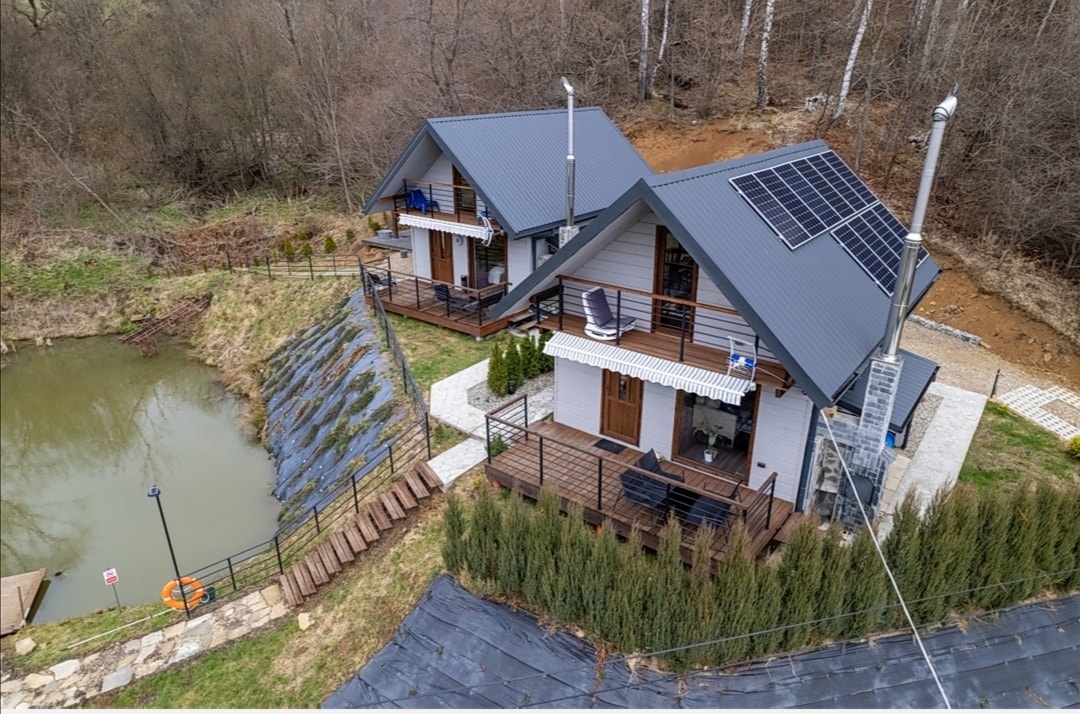 Two modern cabins are situated on a gentle hillside beside a pond. Each cabin features a deck with outdoor seating and is equipped with solar panels on the roof. Pathways lead down to the water's edge, surrounded by natural landscaping and greenery.