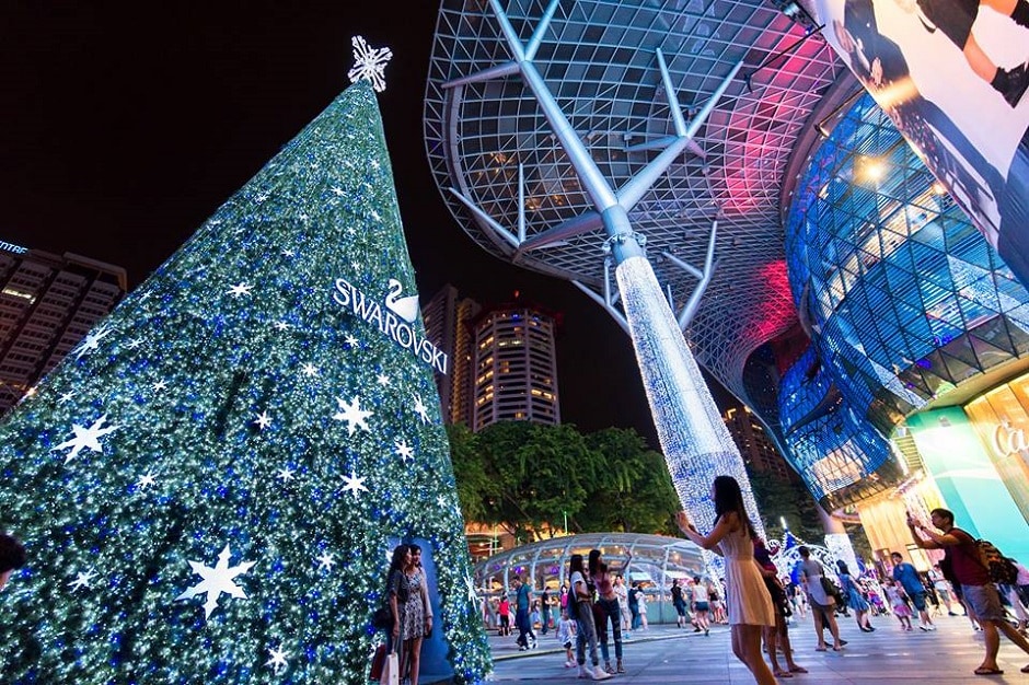 A brightly lit Swarovski Christmas tree stands prominently at night, adorned with sparkling lights and decorative stars. Nearby, busy pedestrians enjoy the festive atmosphere, set against a backdrop of modern architecture and illuminated facades.