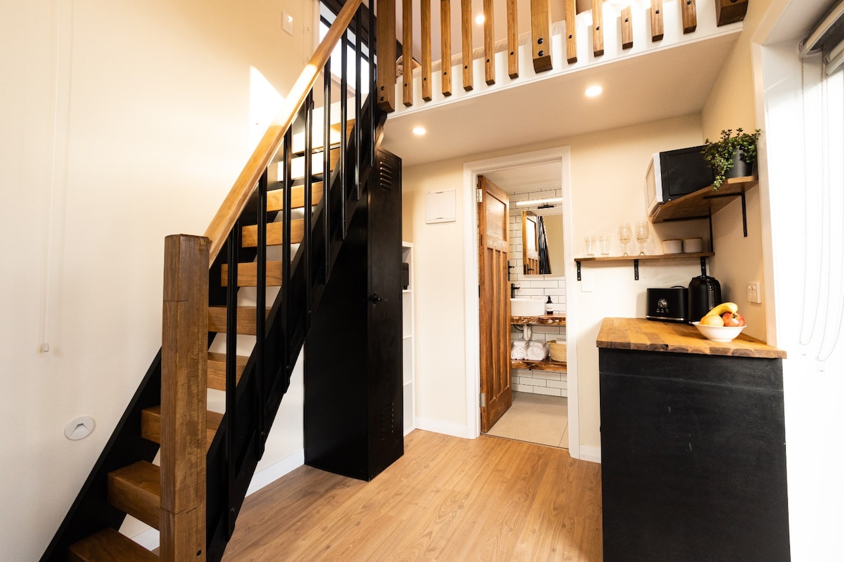 The interior of the tiny house showcases a staircase leading to an upper loft surrounded by wooden railings. A wooden countertop is visible, accompanied by minimal decor. Natural light floods the space through well-placed windows, enhancing the open and airy feel.