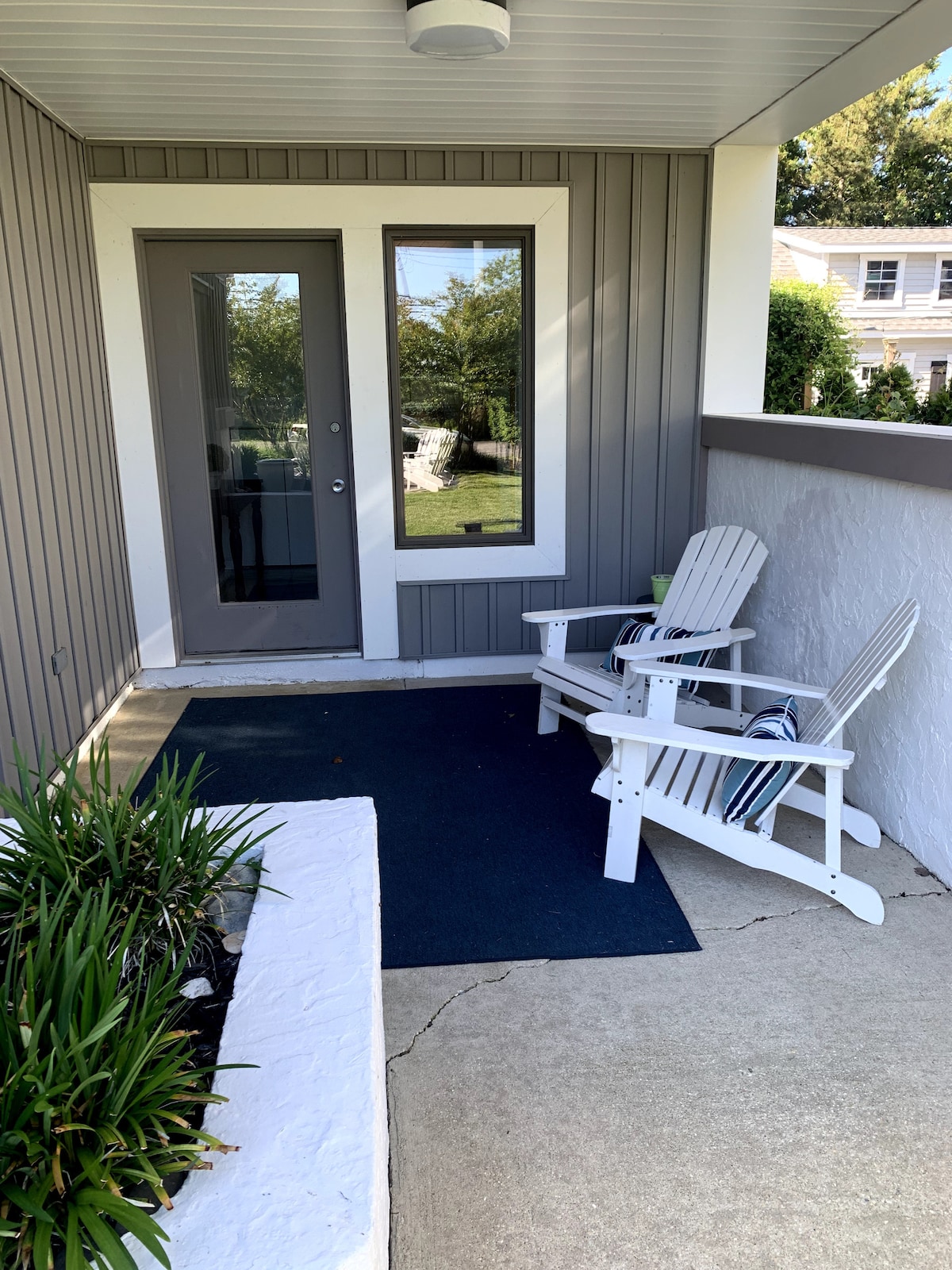 A welcoming porch area features two white Adirondack chairs positioned on a dark rug. A gray door with large windows reflects greenery and sunlight. Lush plants are bordered by a white stone planter, contributing to an inviting entryway.