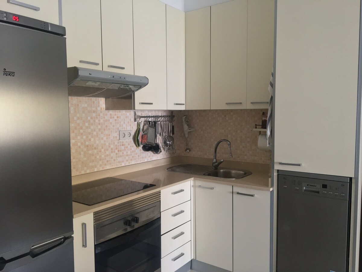 A well-equipped kitchen features modern appliances including a refrigerator, oven, and dishwasher. Light-colored cabinetry surrounds a smooth countertop, with utensils and cooking tools conveniently organized. A small sink is positioned beneath a window, creating a functional cooking space.