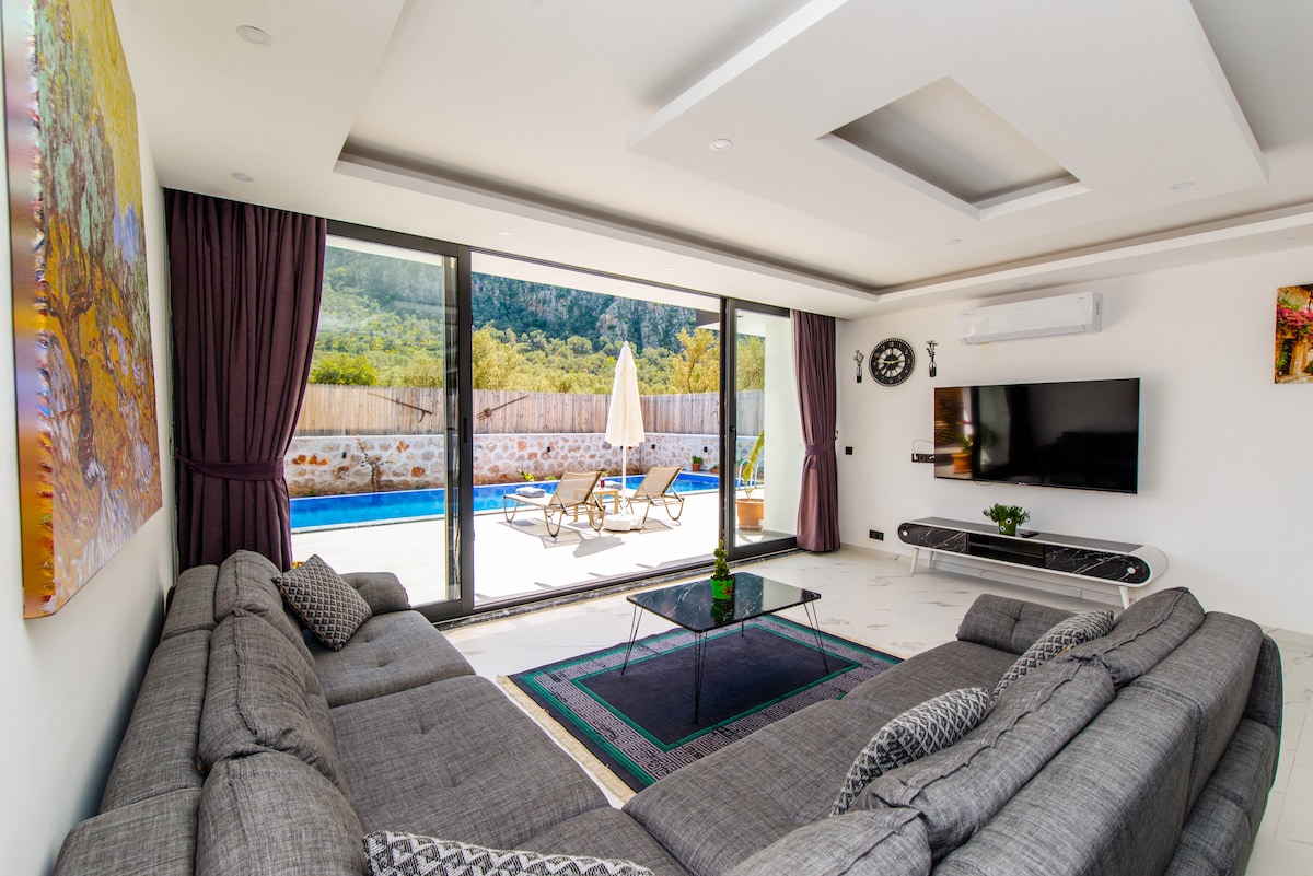A spacious living area is highlighted by large glass doors opening to the outdoor pool. Comfortable grey sofas are arranged around a central coffee table on a patterned rug. Artwork adorns the walls, and natural light fills the space, complemented by green views.