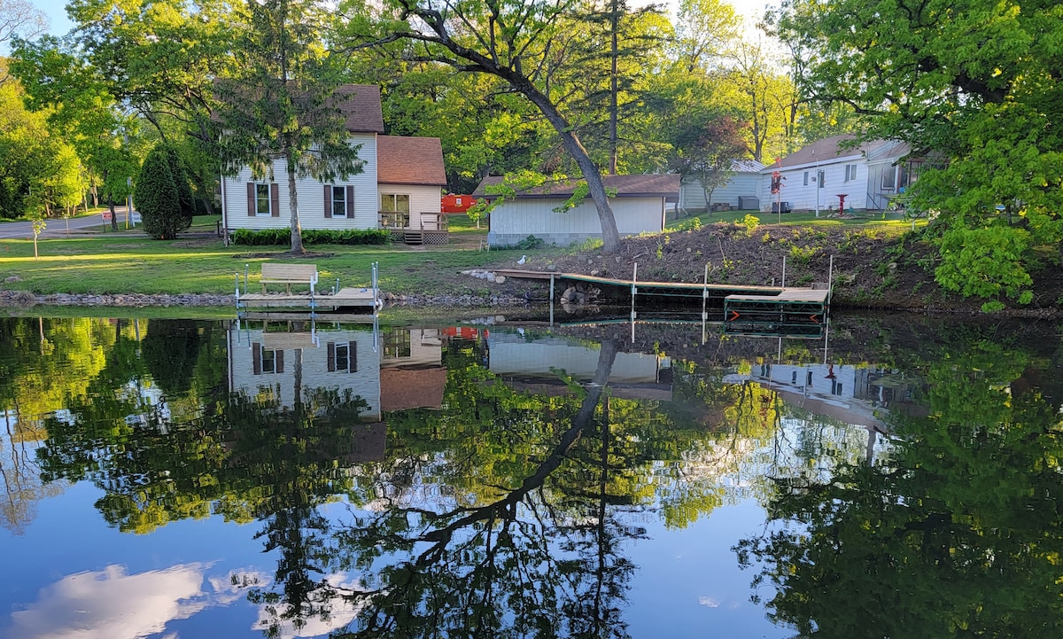 A serene view of a lake reflects trees and a farmhouse. The lush greenery surrounds the property, with a small dock visible at the water's edge. Two neighboring structures are positioned nearby, contributing to the peaceful lakeside atmosphere.