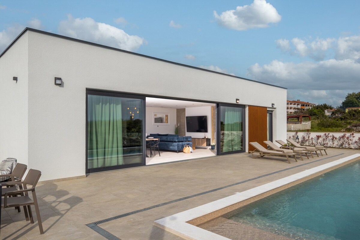 An expansive outdoor terrace is visible, featuring a swimming pool alongside multiple sun loungers. Large glass doors open to a bright living area, inviting natural light. Soft furnishings and modern decor are evident within, while a scenic backdrop can be seen beyond the pool.