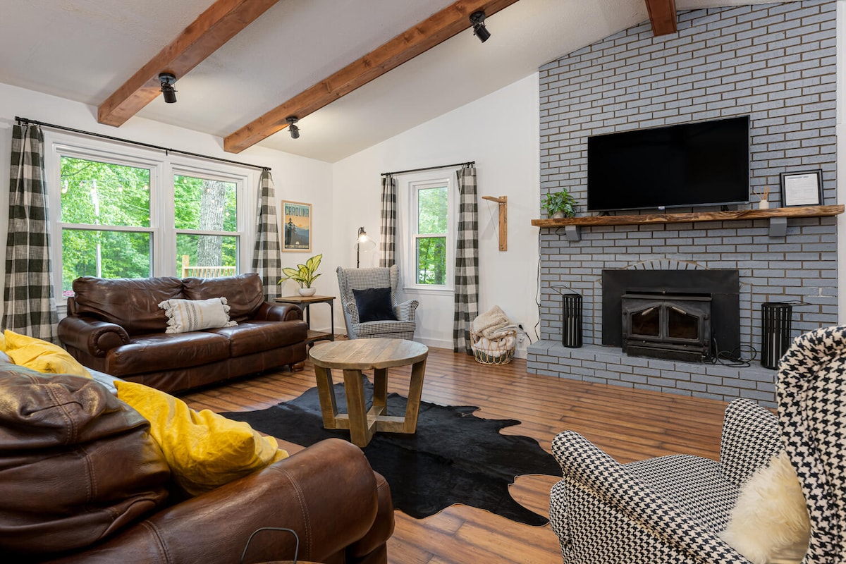The open living area features a cozy seating arrangement comprised of a leather sofa and comfortable armchair, complemented by a wooden coffee table. Natural light filters through crisp curtains, illuminating the rustic fireplace set against a gray brick wall. Wooden beams are visible overhead.