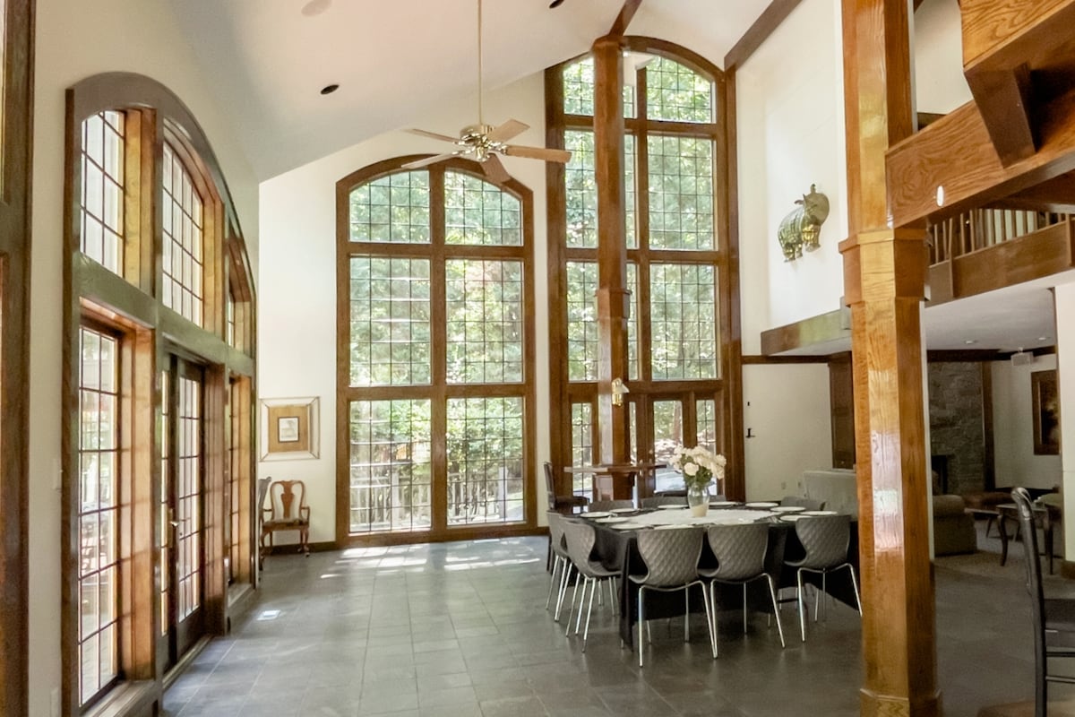 A spacious dining area features large windows that allow natural light to fill the room. A table is set for dining with dark chairs arranged around it. Elegant woodwork frames the space, with high ceilings supported by wooden beams, creating an airy atmosphere.