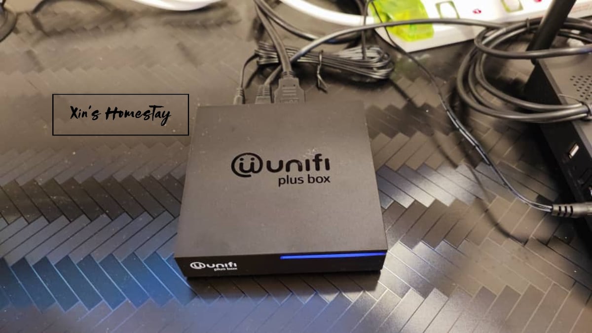 A black Unifi Plus box is positioned on a textured surface with various cables connected. The device features the Unifi logo, offering high-speed internet access. Surrounding elements suggest a functional setup, ideal for guests to stay connected during their visit.