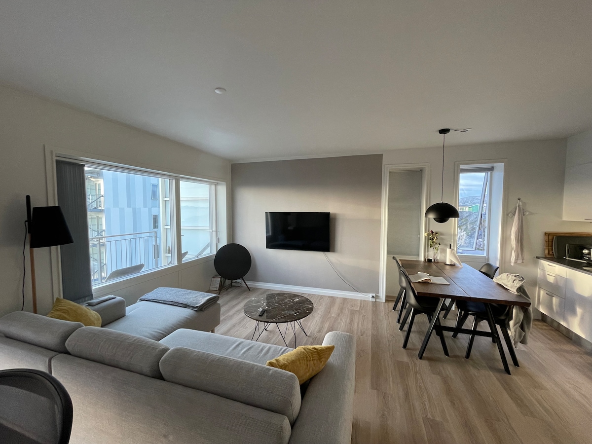 A modern living space features large windows that allow natural light to fill the room. A cozy sofa and a rounded chair are positioned around a glass coffee table. A dining table with seating is visible, along with a mounted television on the wall.