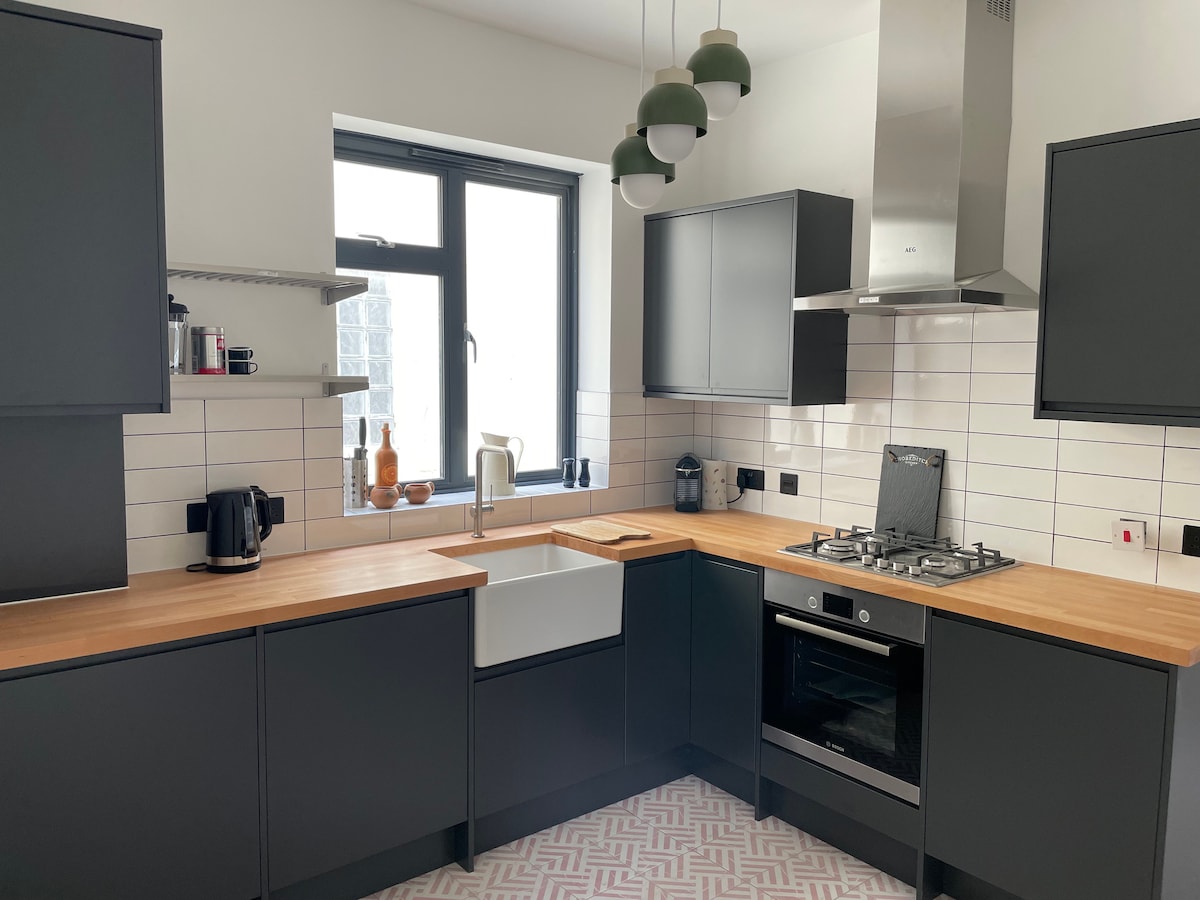 A modern kitchen features dark cabinetry and a light wooden countertop. A farmhouse-style sink is centrally located, with a gas stove and oven nearby. Green pendant lights hang above the workspace, and a window allows natural light to illuminate the area.
