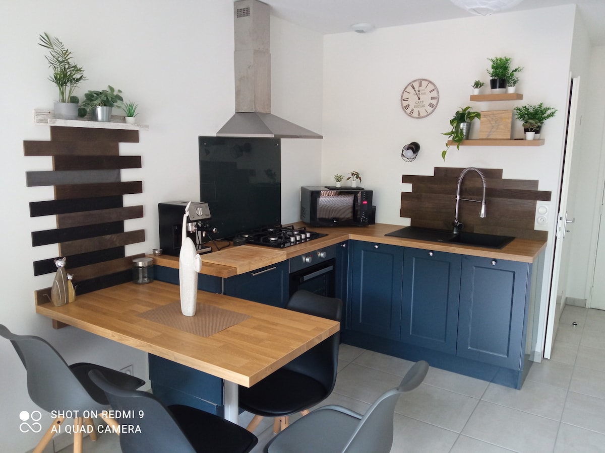 A modern kitchen is presented with dark blue cabinetry and wooden countertops. Essential appliances, including a stovetop and microwave, are visible. Greenery is featured on the shelves, while a dining table with sleek chairs is positioned nearby. A clock is mounted on the wall.