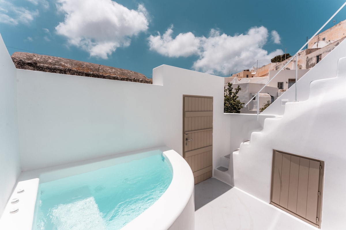 An outdoor area features a round, turquoise pool set against whitewashed walls and clear blue skies. A single door leads to another space, and a staircase ascends to the upper level, framed by gentle clouds overhead.