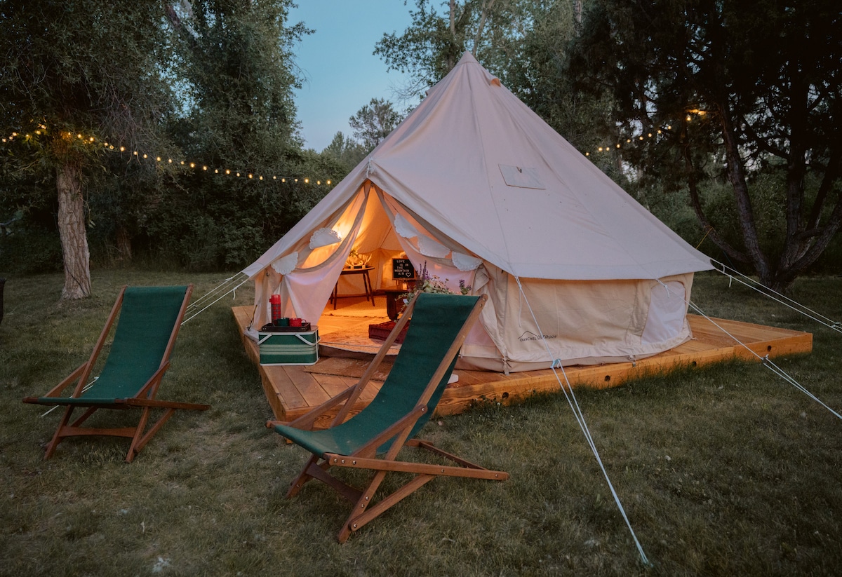 A spacious glamping tent is set on a wooden deck surrounded by trees. Two green lounge chairs are positioned nearby, facing the entrance. Soft lighting is visible above the tent, creating a cozy atmosphere in the evening light.