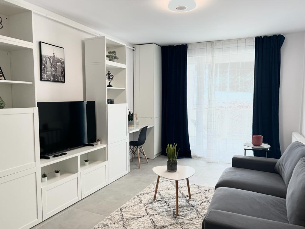 A modern living area features a gray sofa facing a wall-mounted television. A white coffee table is positioned on a patterned rug. Floor-to-ceiling windows provide natural light, adorned with sheer curtains. A small desk with a chair is visible in the background.