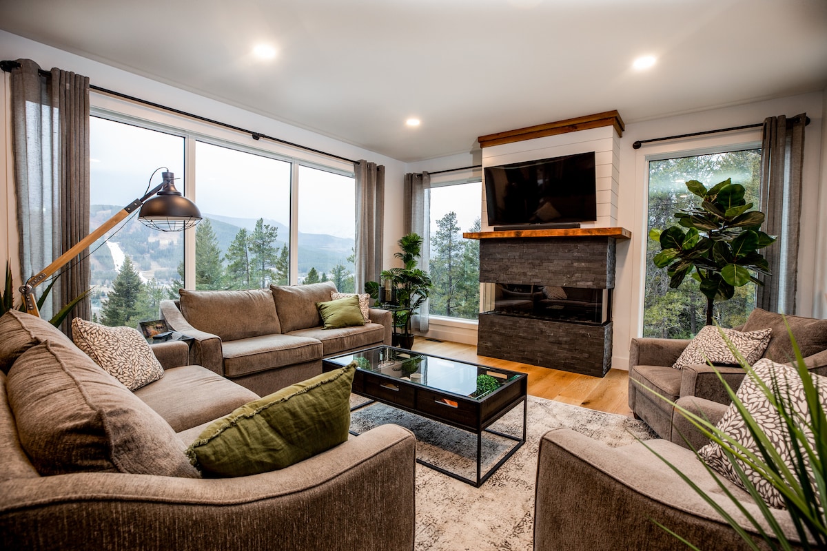 A spacious living room is furnished with a large sectional sofa, accent chairs, and a coffee table. Large windows provide views of the surrounding mountains, while a modern fireplace adds a focal point. Soft lighting enhances the inviting atmosphere.
