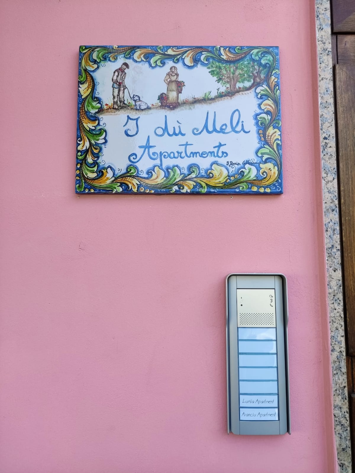 A colorful sign displaying 'I dui Meli Apartments' is mounted on a pink wall, featuring a decorative frame with images of people and nature. Below the sign, an intercom system with multiple buttons is positioned for visitor access.
