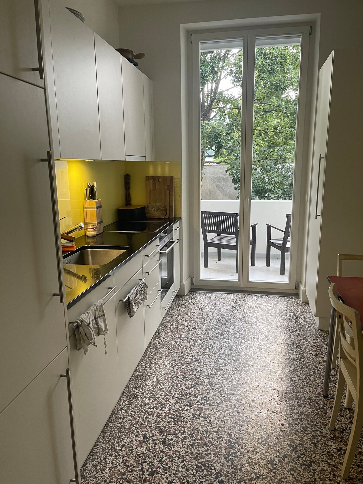 The kitchen features modern appliances with sleek cabinetry and a long countertop. Natural light enters through large glass doors that lead to the balcony. A small dining area with chairs is positioned nearby, while the unique terrazzo flooring adds texture to the space.