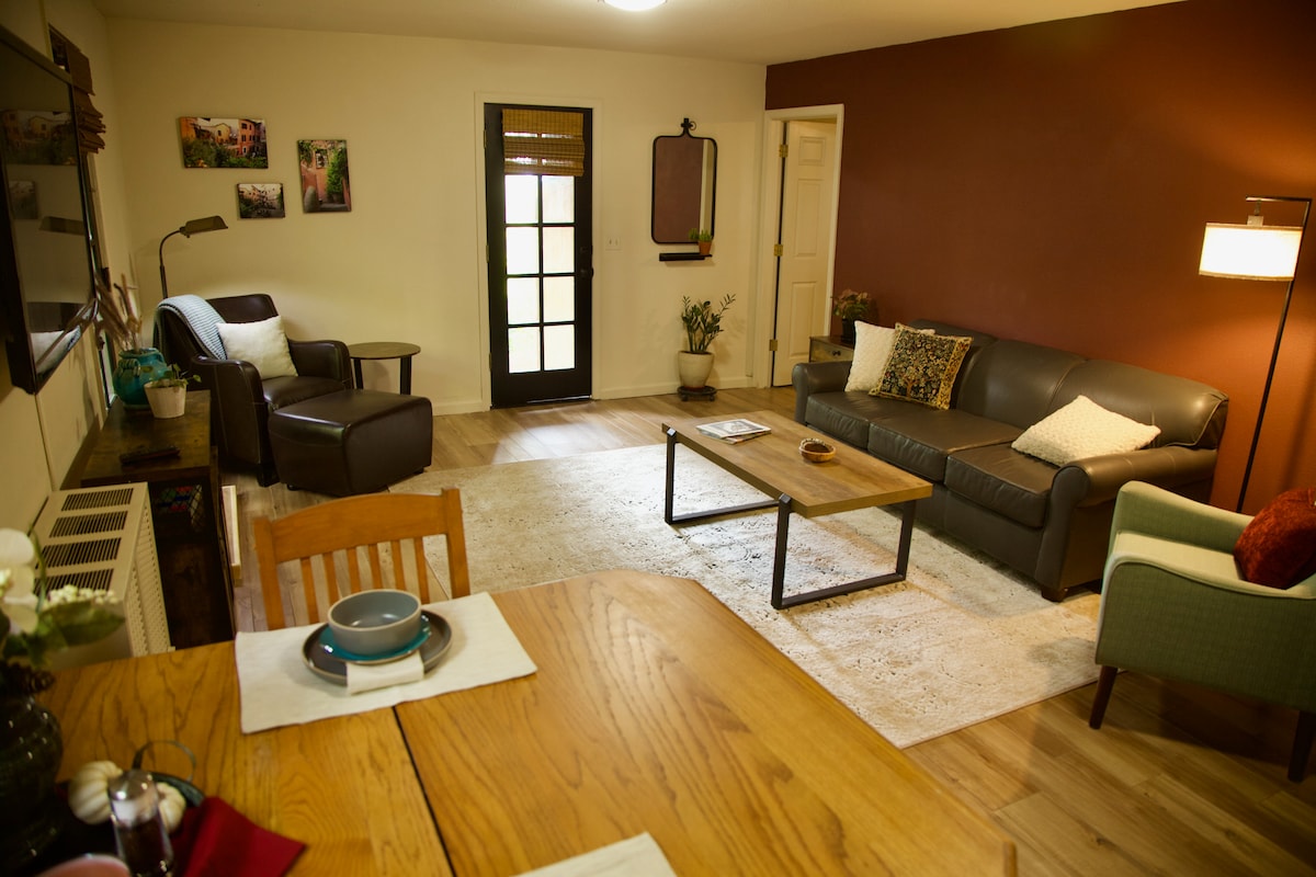 The living area features a leather couch and a reading chair positioned near a wooden dining table set with plates. A coffee table sits on a soft rug, with an entry door leading to the outside. Soft lighting enhances the space's cozy atmosphere.