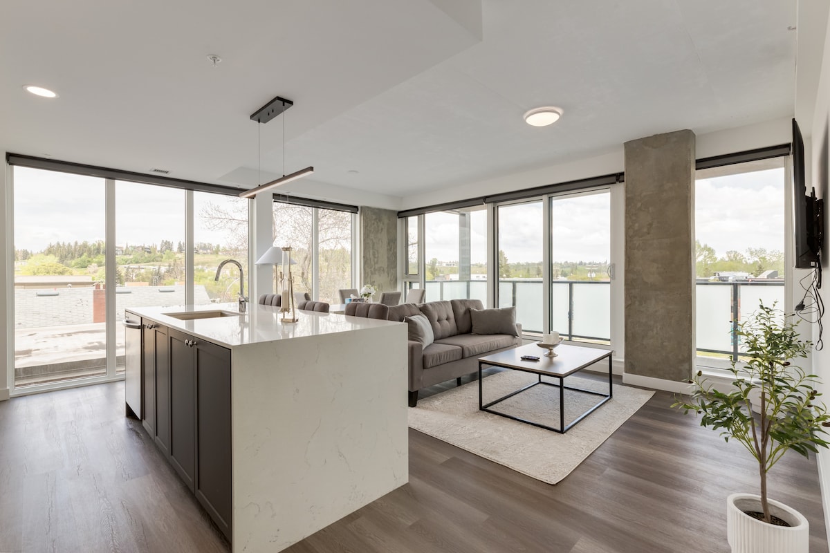 A modern living space is highlighted by large windows allowing natural light to fill the area. The open layout showcases a comfortable seating arrangement, a sleek kitchen island with a marble surface, and a spacious area rug. A peaceful view of greenery is visible through the glass doors.