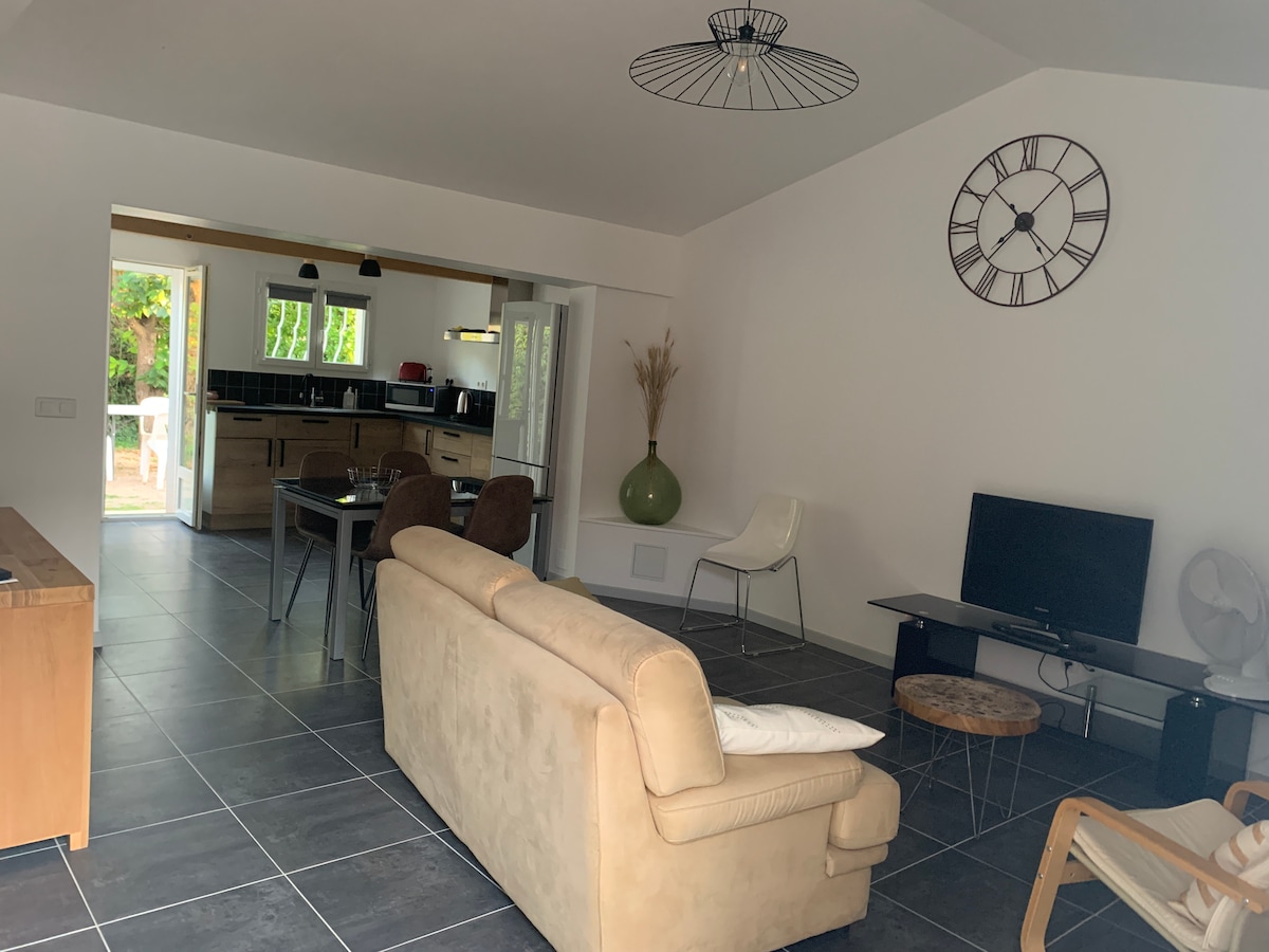 A spacious living area is adorned with slate flooring, featuring a comfortable beige sofa and a wooden sideboard. An entertainment unit supports a television, while a dining table is set for four. Natural light filters through the open doorway leading to the outdoor space.
