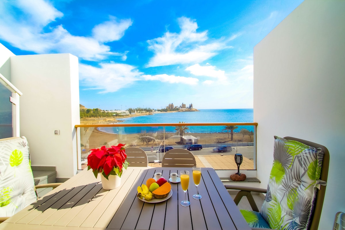 A spacious terrace features a wooden dining table set for two, overlooking the ocean. Bright tropical plants and a bowl of fresh fruit are displayed on the table, with two drinks accompanying them. The coastline and beach can be seen in the background under a clear blue sky.