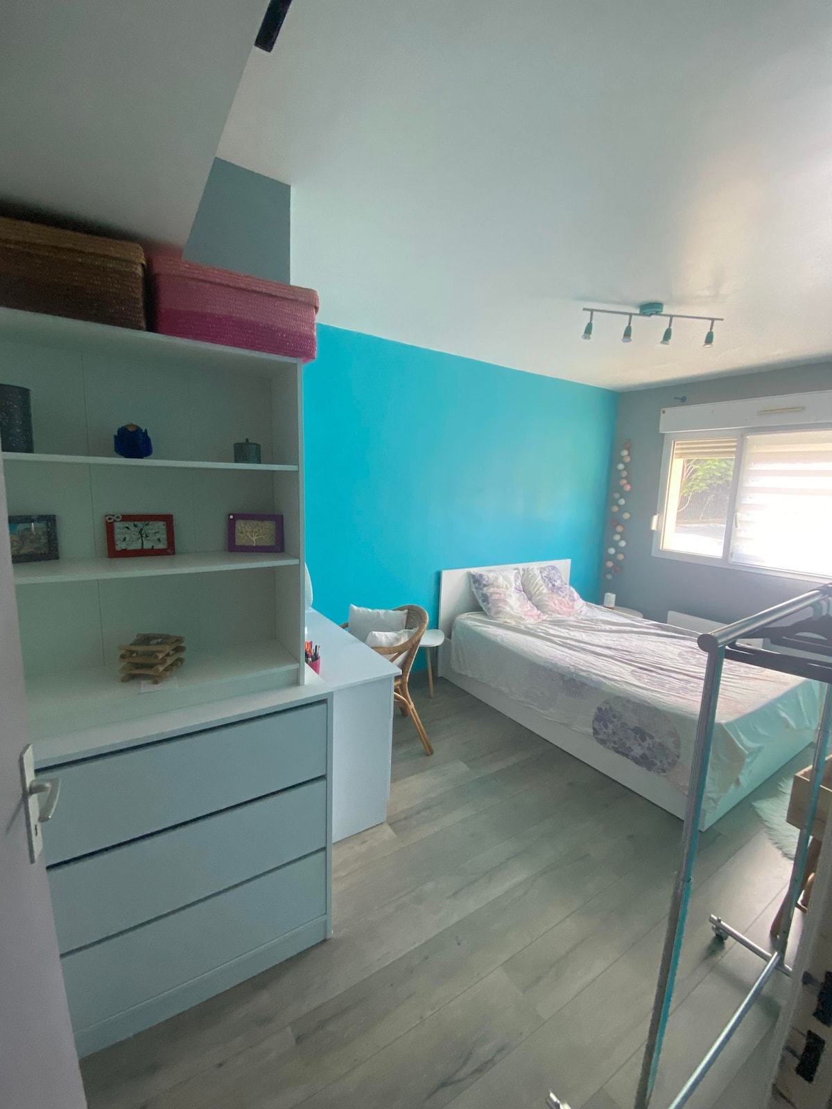 A bright bedroom features a spacious bed adorned with light bedding. A vibrant blue wall adds color to the room, while a white shelving unit and a small chair are positioned nearby. Natural light enters through a window with partially closed blinds.