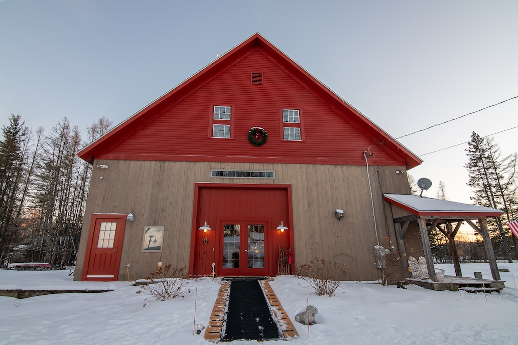 The exterior of a rustic lodge features a striking red roof and wooden siding, framed by snowy grounds. Two symmetrical entry doors welcome guests, with a wreath adorning the center above the main entrance. Surrounding trees stand tall in the background.