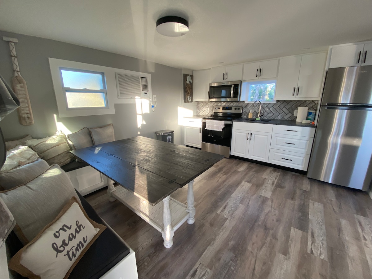 An open kitchen area is shown, featuring a large wooden table surrounded by comfortable seating. White cabinetry complements stainless steel appliances, while natural light enters through a window. Textured flooring adds warmth, and a decorative pillow with the phrase 'on beach time' is visible on the sofa.