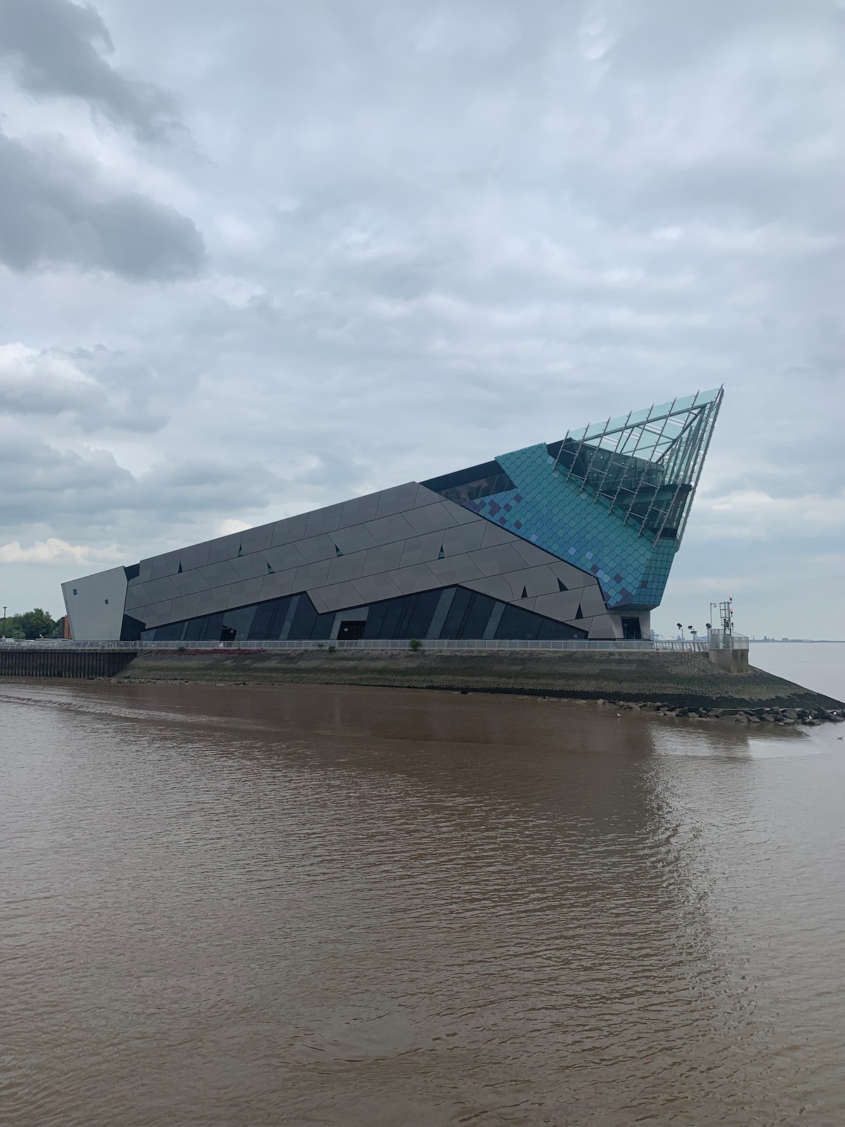 A distinctive modern building is situated at the water's edge, featuring an angular design with a glass pyramid peak. Its geometric façade reflects a contemporary architectural style, complemented by the surrounding water and cloudy sky.