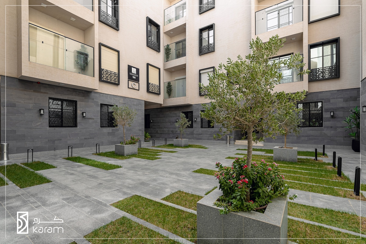 A central courtyard is presented, featuring neatly arranged green grass and decorative plants. Gray stone walkways intersect the space, framed by the surrounding building's modern architecture. Natural light filters into the area, enhancing the inviting ambiance of the outdoor setting.