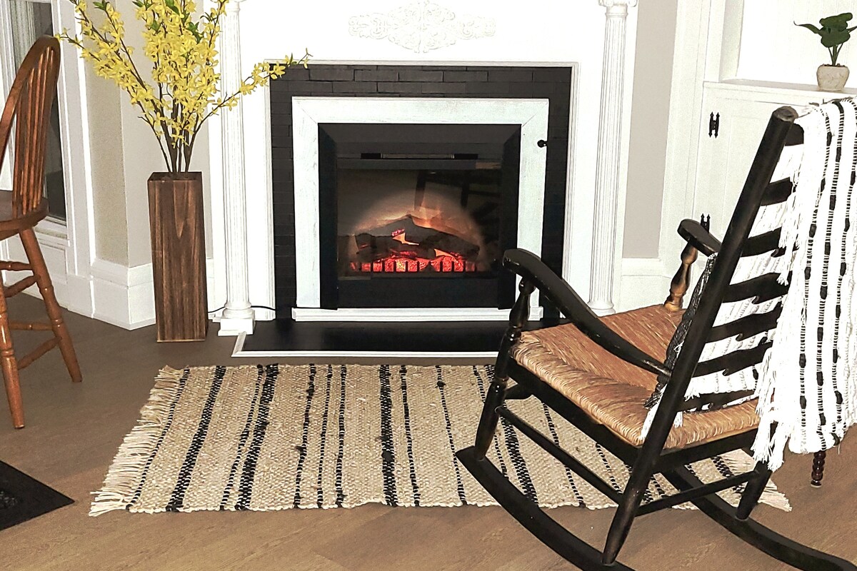 A cozy corner of the living room features a black electric fireplace surrounded by a sleek white mantel. A wooden rocker sits beside a textured area rug, while yellow floral arrangements in a vase add a touch of warmth to the space.